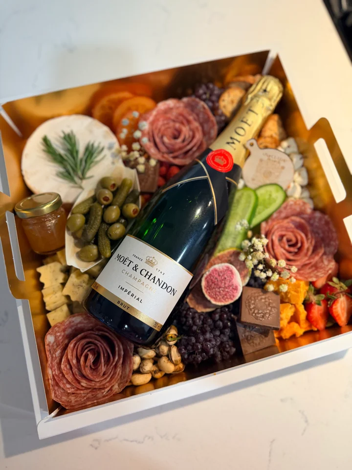 A charcuterie gift box arranged with fruit, cheese, florals, and a champagne bottle