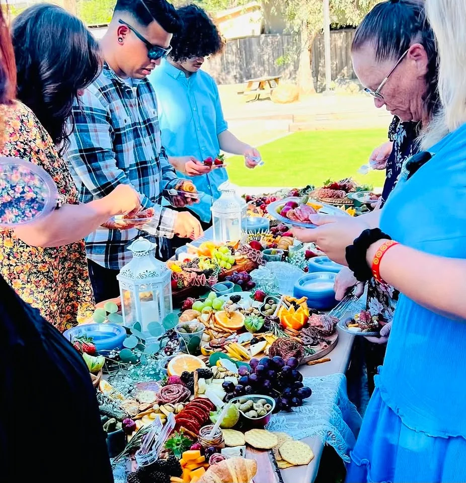 Guests gathering around a generously styled outdoor charcuterie table with fruit, cheese, crackers, and cured meats