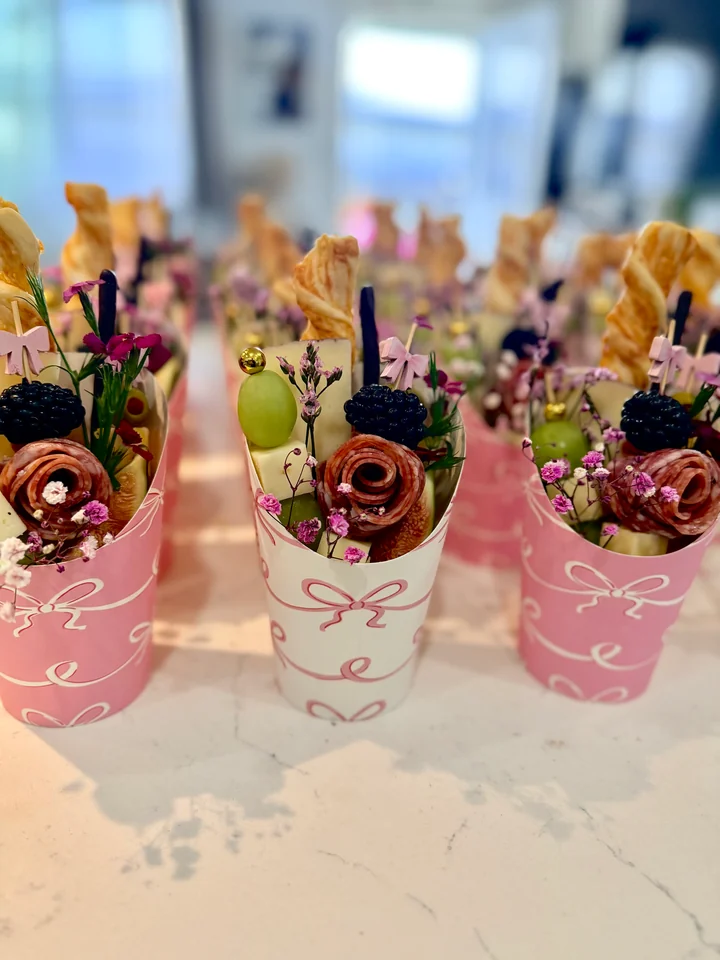 Pink bow charcuterie cups filled with salami roses, berries, flowers, cheese, grapes, and crackers