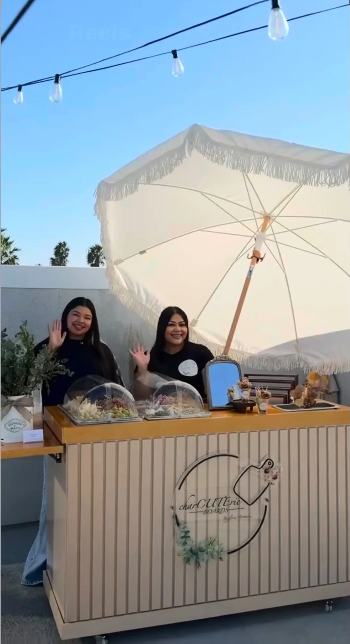 Two team members waving behind an outdoor charcuterie cart beneath a fringed umbrella and string lights